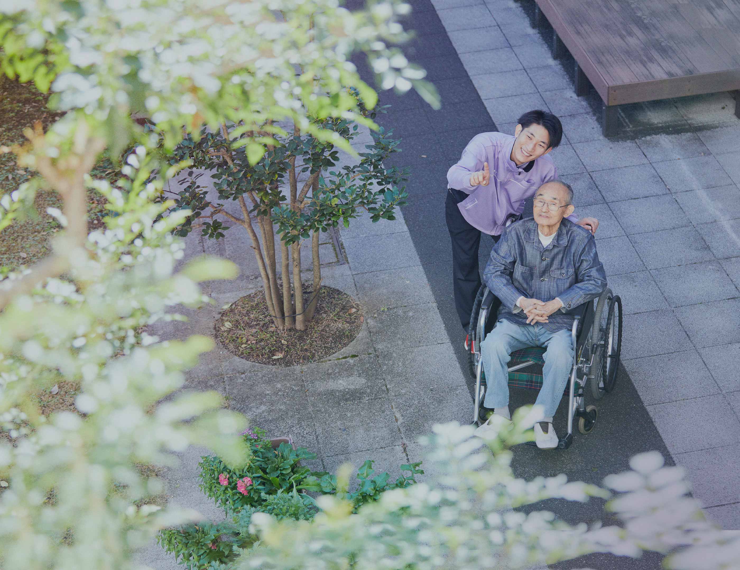 職員が車椅子利用者を庭園内で介助している様子。季節の花々が咲く環境での入居者の生活場面。