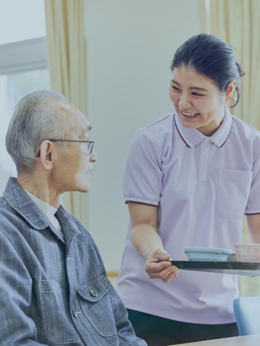 介護職員が高齢入居者に笑顔で対応する様子。居室内での日常的なケアシーン。